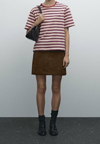 Henkilö, joka käyttää lyhythihaista raidallista paitaa, ruskeaa hametta, mustia sukkia ja mustia kenkiä, pitäen pientä mustaa käsilaukkua olkapäällä.