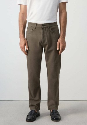 Homme portant un pantalon droit vert olive, une chemise blanche à manches courtes et des mocassins noirs, debout sur un sol gris devant un fond blanc.