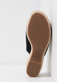 Semelle d'une espadrille noire avec une semelle extérieure en caoutchouc texturé et une semelle intermédiaire enveloppée de corde, posée en angle contre un fond blanc d'angle.