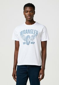 Camiseta de algodón blanca con un gráfico azul de "WRANGLER" con alas y el año "1947". Cuello redondo estándar y mangas cortas.