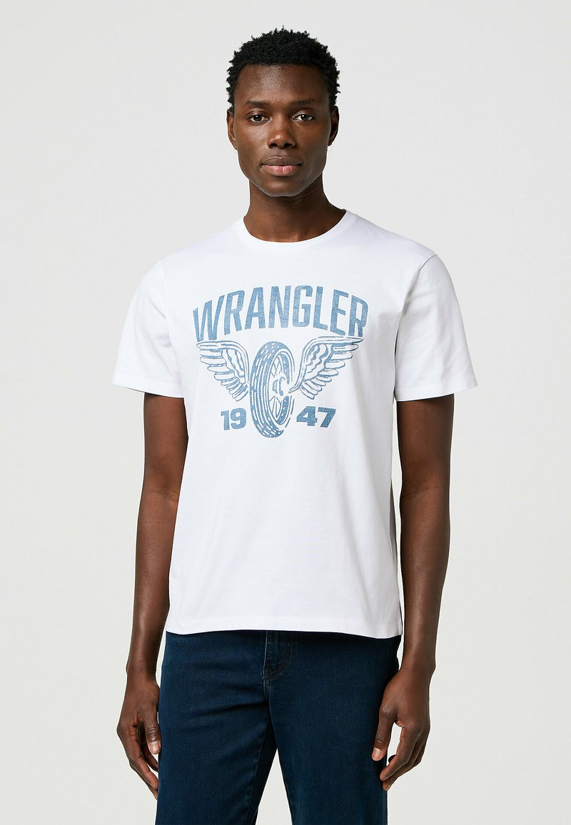 Camiseta de algodón blanca con un gráfico azul de "WRANGLER" con alas y el año "1947". Cuello redondo estándar y mangas cortas.