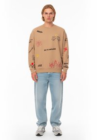 Felpa beige con vari design grafici, tra cui rose e fiamme. Abbinata a jeans blu chiaro a gamba dritta e sneakers grigie sportive.