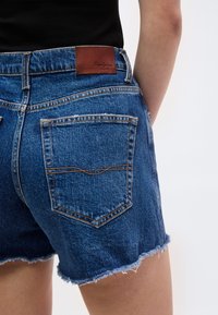 Κοντινό πλάνο ατόμου που φοράει μπλε τζιν σορτς με ξεφτισμένες άκρες και καφέ δερμάτινο ετικέτα με το λογότυπο Pepe Jeans στη μέση.