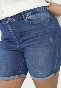 Tamsiai mėlyni džinsiniai šortai, pasižymintys suvyniotu kraštu ir subtiliais dėvėjimosi požymiais. Įrengti priekiniai kišenės ir metalinė mygtuko uždarymo sistema.