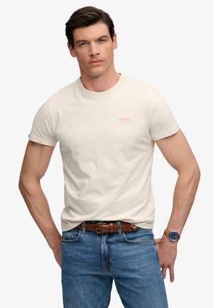 Mężczyzna ubrany w kremową koszulkę Superdry, niebieskie dżinsy z brązowym plecionym paskiem i zegarek na lewym nadgarstku.