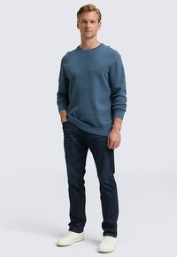 Mėlynas megztinis su apvalia iškirpte, derinamas su tamsių džinsų kelnėmis ir baltomis sportinėmis avalynėmis. Modelis stovi su rankomis kišenėse priešais paprastą foną.