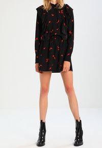 Robe noire à motif de feuilles rouges, épaules froncées, taille élastique et manches longues. Portée avec des bottines noires brillantes.