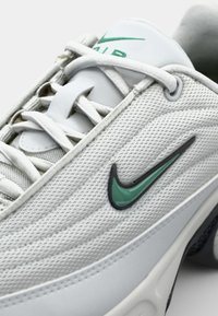 Valge spordijalats, millel on tekstuuriga pehme ülaosa, rohelise Nike swoosh logo ja mustad aksendid. Ketsikummud on asetatud keele peale.