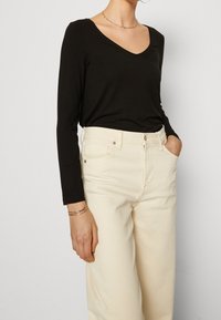 Maglietta nera a maniche lunghe con scollo a V abbinata a pantaloni beige a vita alta. La maglietta presenta un tessuto liscio, mentre i pantaloni hanno una finitura strutturata.