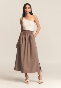 Top de canalé beige claro con un solo hombro, combinado con una falda larga de lino marrón que presenta una cinturilla elástica y una abertura lateral. Se lleva con zapatos de alpargata trenzados.