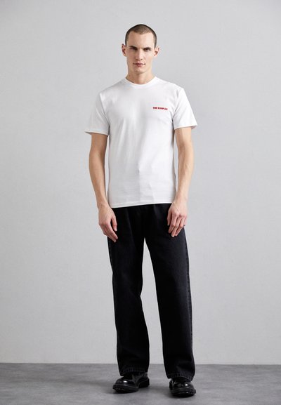 Camiseta blanca de algodón con mangas cortas, que presenta un logo de texto rojo en el lado izquierdo del pecho. Se lleva con pantalones negros holgados y zapatos negros.