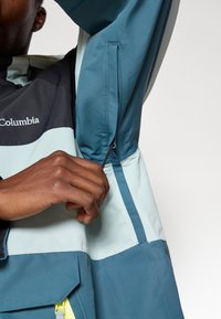 Casaco à prova de água em verde-água e preto, com um bolso com zíper, punhos ajustáveis e blocos de cor contrastantes. Textura suave.