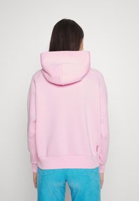 En person med långt hår bär en rosa hoodie och blå kordflätsbyxor, sedd bakifrån mot en enfärgad vit bakgrund.