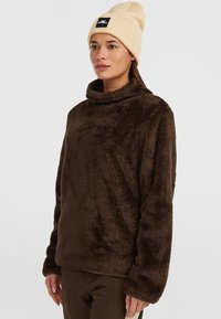 Brun, faux fur pullover med høj krave og lange ærmer. Båret med en beige strikket hue, der har et sort logo-patch.
