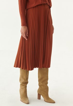 Person trägt einen rostfarbenen plissierten Midirock, einen rostfarbenen Pullover und beige kniehohe Wildleder-Stiefel mit Absatz auf weißem Hintergrund.