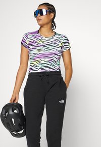 Tæt siddende skjorte med multikoloreret zebra-print og korte ærmer, parret med sorte sweatpants med et logo, og en sort hjelm holdt i den ene hånd.