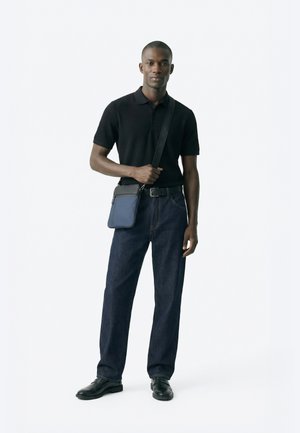 Άνδρας που φοράει μαύρο πόλο πουκάμισο, σκουρόχρωμο τζιν, μαύρα παπούτσια και μπλε τσάντα χιαστί με ρυθμιζόμενο ιμάντα, μπροστά σε απλό λευκό φόντο.