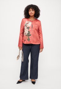 Giacca di raso corallo con stampa floreale a sinistra, scollatura rotonda, maniche lunghe, abbinata a pantaloni in denim scuro e scarpe nere.