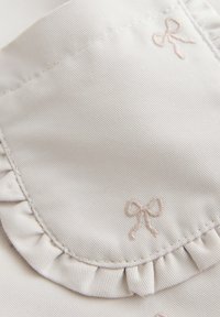 Primer plano de tela beige claro con lazos cosidos y un detalle de borde fruncido en una prenda.