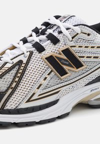 Gros plan d'une chaussure de sport blanche et noire avec une tige en mesh, des détails noirs et dorés, des lacets blancs et le logo "N Lock" sur le côté.