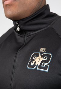 Zwarte ritsjack met een hoge kraag. Bevat een grafisch ontwerp met "82" en een illustratie. Zilveren ritstrekkertje voegt detail toe.