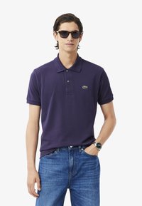 Ung man som bär solglasögon, en lila polo med en logotyp och blå jeans, står med händerna i fickorna mot en enhetlig bakgrund.