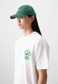 Cappello in cotone verde con visiera curva e testo ricamato. Indossato con una t-shirt bianca con una stampa grafica colorata sul lato sinistro del petto.