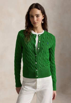 Mujer de pie frente a un fondo neutro, vistiendo un cárdigan verde de punto grueso sobre una blusa blanca y pantalones blancos, con pendientes visibles.