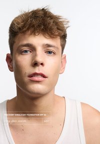 Teint clair, yeux bleus, cheveux bruns courts et ébouriffés, expression neutre, portant un débardeur blanc. Étiquette du produit : "LE FOND DE TEINT TOUTES LES JOURS SPF 50."