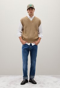 Gilet lavorato a maglia beige sopra una camicia bianca con bottoni, abbinato a jeans blu arrotolati e scarpe nere, più un cappellino con logo verde.