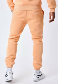 Pantalones de chándal color durazno en tejido suave, con puños elásticos en los tobillos y bolsillos laterales. Llevados con zapatillas Nike blancas y calcetines grises.