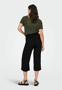 Haut vert en tricot à manches courtes, associé à un pantalon noir raccourci à taille élastique. Le mannequin porte des sandales noires. Design et textures simples.