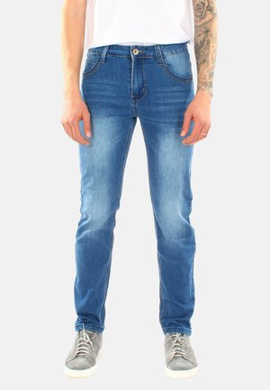Uomo con jeans slim fit blu e sneakers grigie, in piedi davanti a uno sfondo bianco semplice, con l'avambraccio destro tatuato visibile.