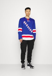 Tricou de hochei albastru cu "RANGERS" în roșu, dungi albe pe mâneci, asortat cu pantaloni cargo negri și adidași negri.