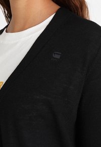 Svart kavaj med V-ringning, tillverkad av ett lätt material. Har en liten varumärkeslogotyp på höger sida.