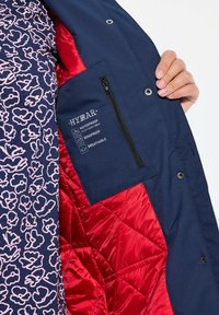 Giacca impermeabile con fodera trapuntata rossa, esterno blu navy e motivo floreale interno rosa. Include una tasca con zip e dettagli del marchio.