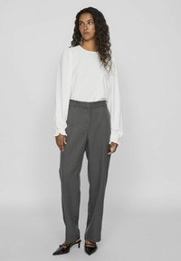 Blusa blanca de manga larga con puños con volantes, combinada con pantalones grises de pierna recta de cintura alta. Unos zapatos negros de tacón completan el conjunto.