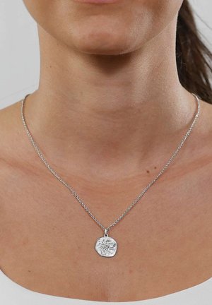 Close-up of a person's neck wearing a silver chain necklace with an irregular round pendant featuring an embossed design.