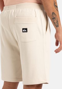 Pantalones cortos beige hechos de una tela suave, con una cinturilla elástica, un bolsillo trasero y un parche de logo negro en el bolsillo.