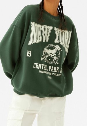 Persoon in een donkergroene trui met "NEW YORK," een hondenafbeelding en witte broek, getoond van nek tot heupen tegen een witte achtergrond.