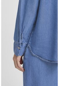Camicia di jeans in azzurro chiaro, con polsini abbottonati e un design a pieghe dalla consistenza morbida. L'orlo è leggermente curvo, rivelando i dettagli della cucitura.