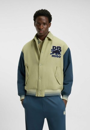 Green and navy bomber jacket with embroidered logo, snap closure, two front pockets, and striped ribbed cuffs and hem. Smooth fabric texture.