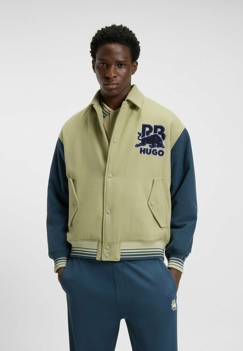 Chaqueta bomber verde y azul marino con logo bordado, cierre de botones a presión, dos bolsillos frontales y puños y dobladillo de canalé a rayas. Textura de tela suave.