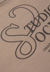Hnědá látka má velký černý grafický textový design, který zobrazuje slova "Studio" a "Společnost" s hladkými, zaoblenými písmeny a akcenty ve stylu popisovače.