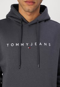 Hoodie gris foncé en mélange de coton, présentant un grand logo blanc "TOMMYJEANS" et un petit emblème tricolore sur le côté gauche.