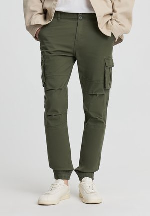 Uomo che indossa pantaloni cargo verde oliva con polsini elasticizzati e sneakers bianche, mani nelle tasche, giacca beige parzialmente visibile.
