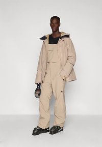 Jonge man in een beige skikostuum en jas, met een skihelm in zijn hand, staande op skischoenen tegen een effen achtergrond.