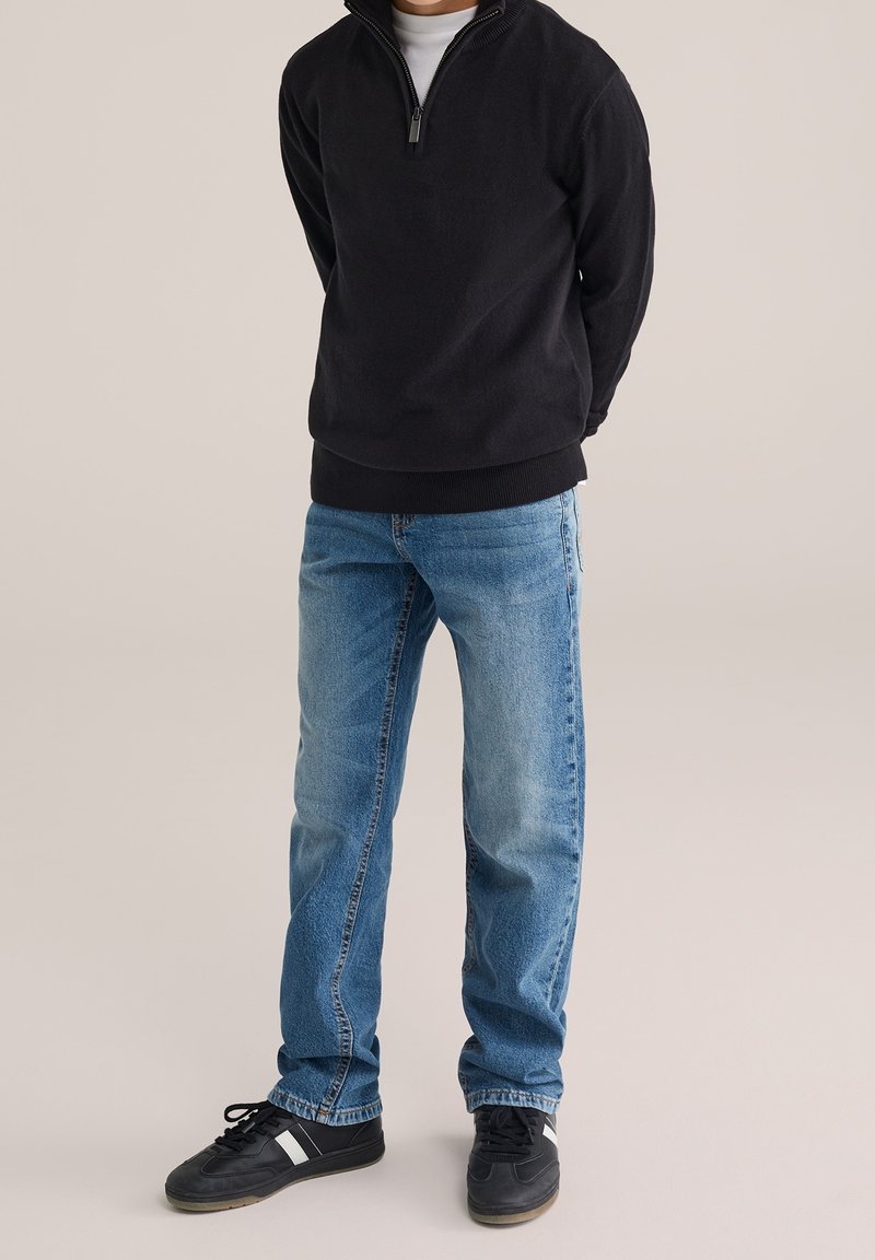 Homme portant un pull noir à fermeture éclair quart, un jean bleu clair et des baskets noires, debout devant un fond uni.