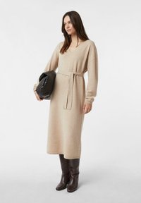 Beżowa dzianinowa sukienka z dekoltem w serek, długimi rękawami, wiązana w talii i z prążkowanym dołem. Do kompletu teksturowana czarna torebka na ramię i brązowe kozaki za kolano.