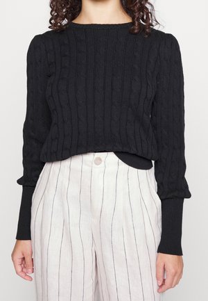 Svart stickad tröja med texturerat flätmönster, långa ärmar och ribbade muddar, kombinerad med ljusa randiga byxor.